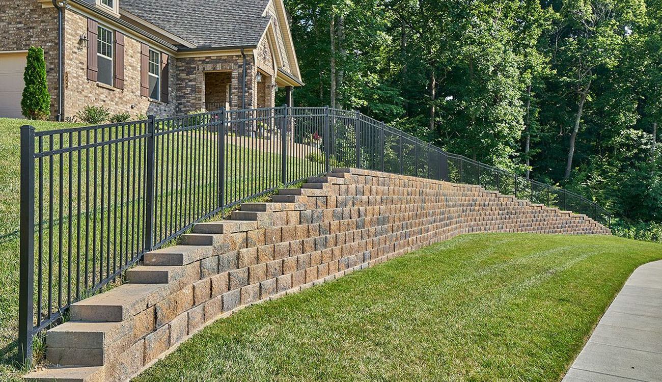 Stone retaining wall ideas with a long tiered wall and metal fence to manage a steep yard and add structure to the landscape.