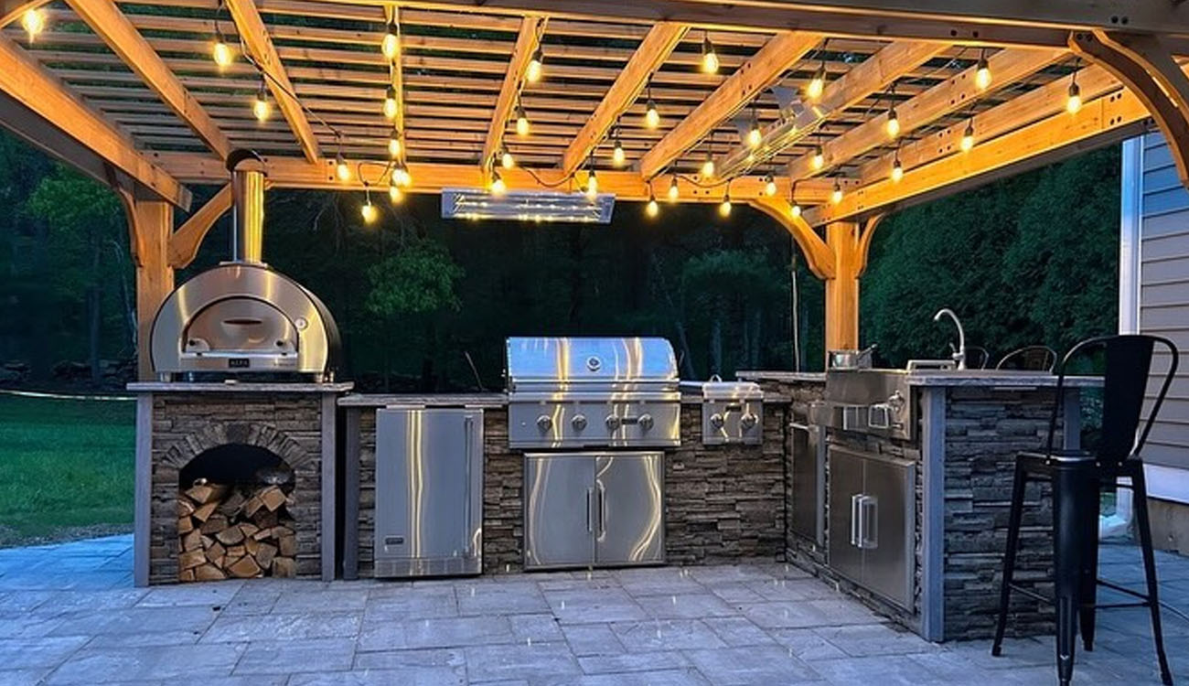 Holistic Patio Hardscaping Services in Fort Worth, TX - Holistic Patio Modern Outdoor Kitchen Ideas A wooden pergola with string lights covers a stone kitchen featuring a premium pizza oven.