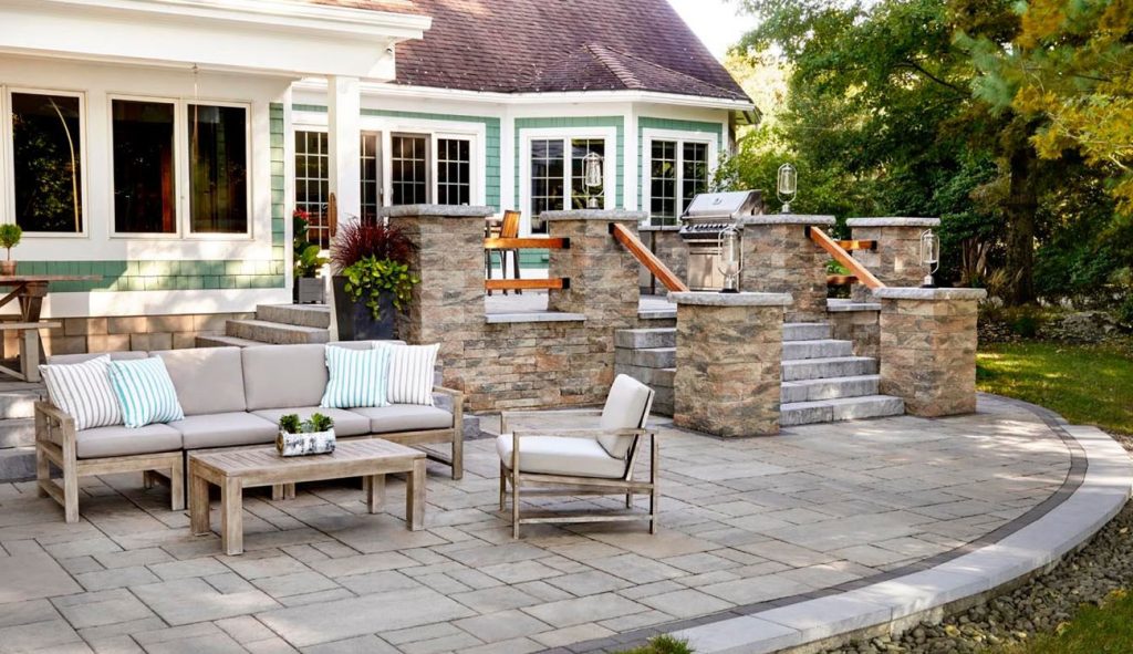 Elegant stone patio includes outdoor kitchen, tiered steps, and comfortable seating beside a bright home.