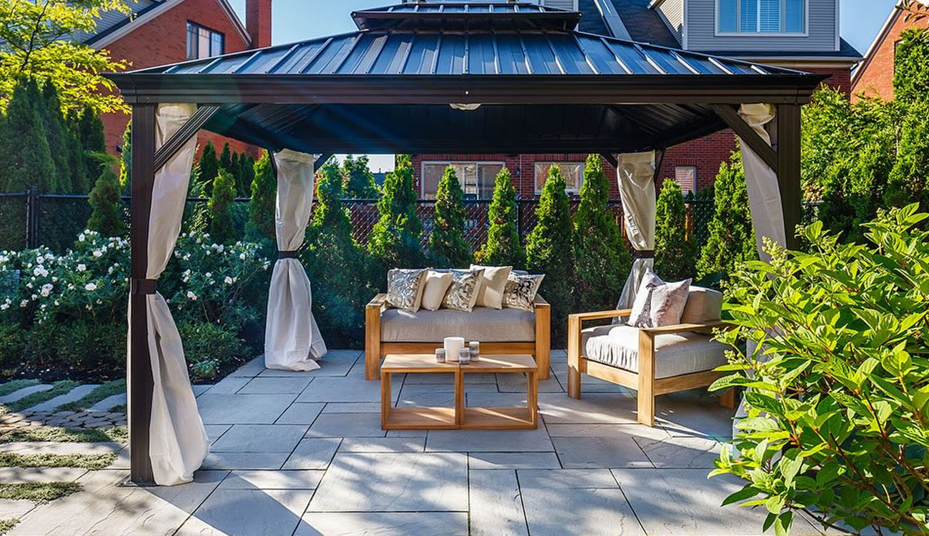 Modern gazebo covers a cozy seating area with stone pavers, soft cushions, and lush garden privacy.